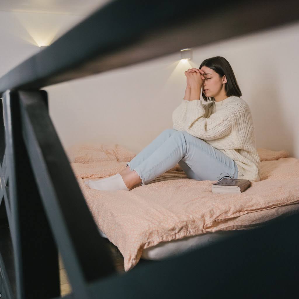 Temos diálogo com Deus na oração. Mulher sentada na cama em posição de estar fazendo uma prece, oração, com Bíblia ao lado. Um óculos esta em cima da Bíblia.